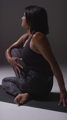 Vertical-Video-Studio-Shot-Of-Mature-Woman-Wearing-Gym-Fitness-Clothing-Sitting-On-Floor-Doing-Pilates-Stretching-Exercises